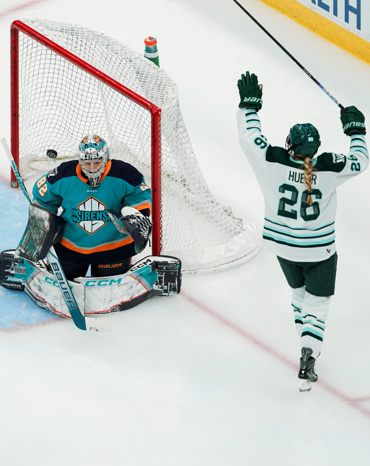 Huber raises her arms in celebration as she skates around the back of the net. She is wearing a white away uniform, while New York's Kayle Osborne is in her crease wearing a teal home one.