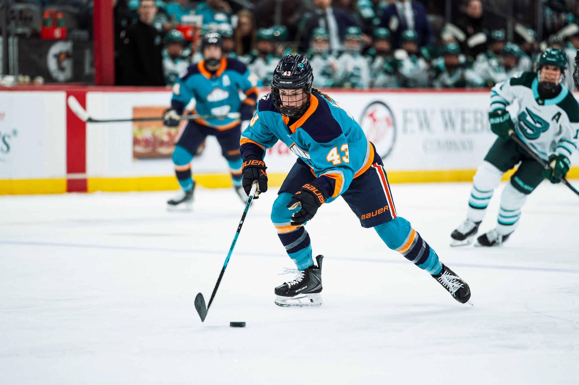 O'Neill looks down as she skates into the offensive zone with the puck in front of her. She is wearing a teal away uniform.