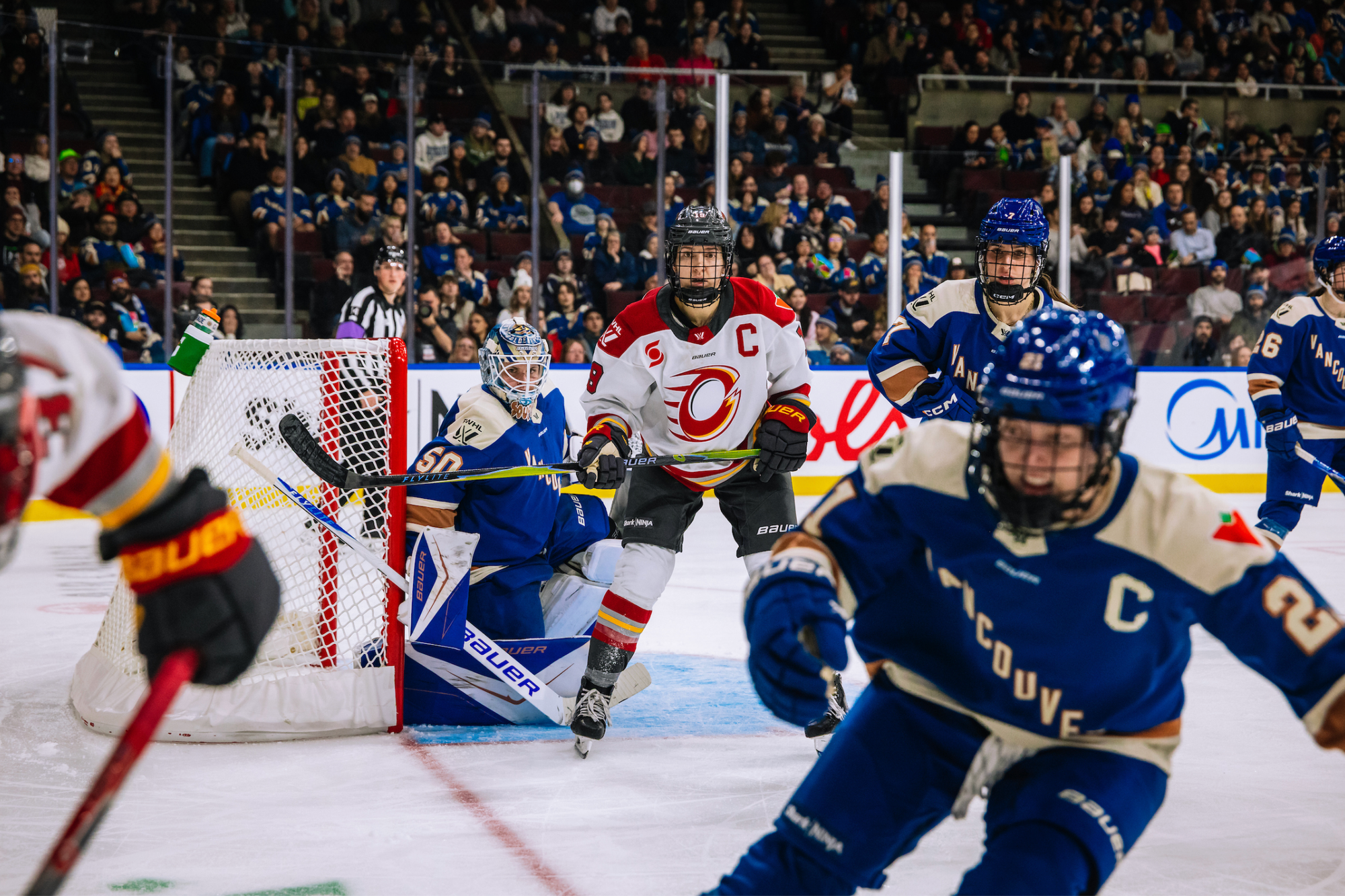 Jenner stands in front of Kristen Campbell's crease with her stick in her hands. She's looking toward the corner at a play unfolding out of frame. Several Vancouver players are in the frame defending. Jenner is wearing a white away uniform, while the Vancouver players are in a blue home one..