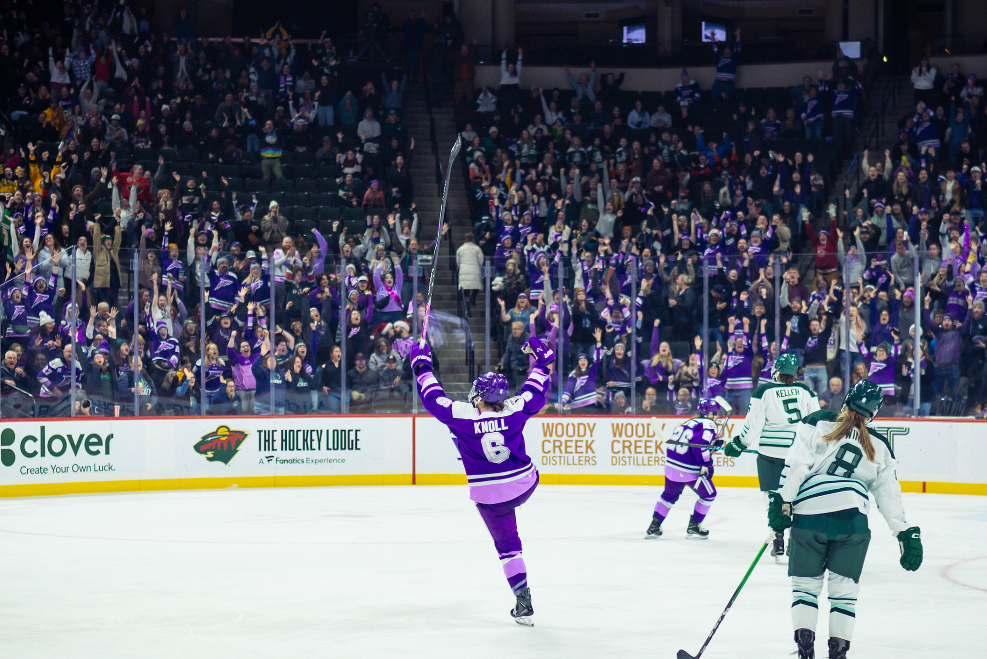 A from-behind shot of Knoll raising her arms and right leg in celebration. The crowd is in the background cheering. Knoll and many crowd members are wearing purple home jerseys. 