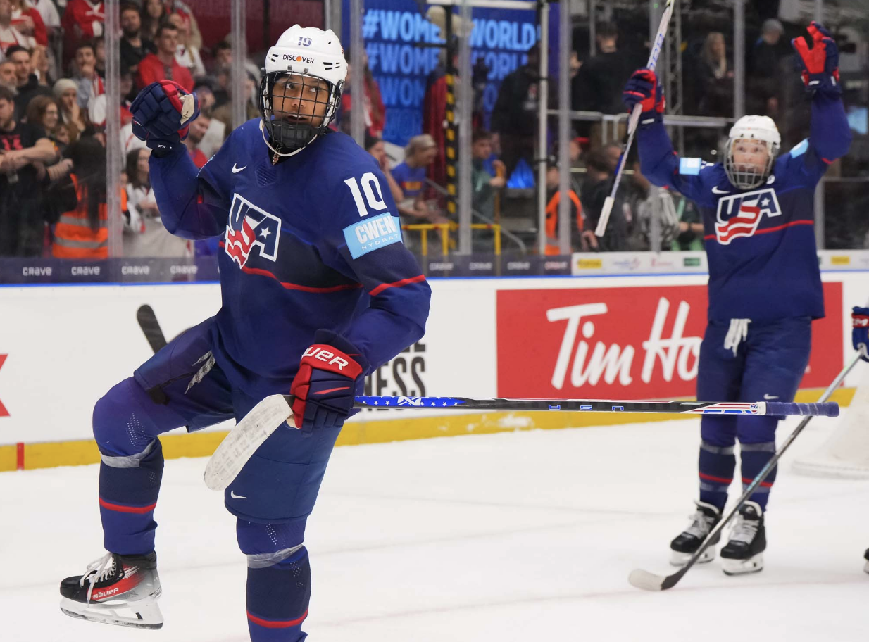 Edwards pumps her first in celebration of her goal, while Hilary Knight raises her arms in celebration in the background. They are both wearing blue home uniforms.