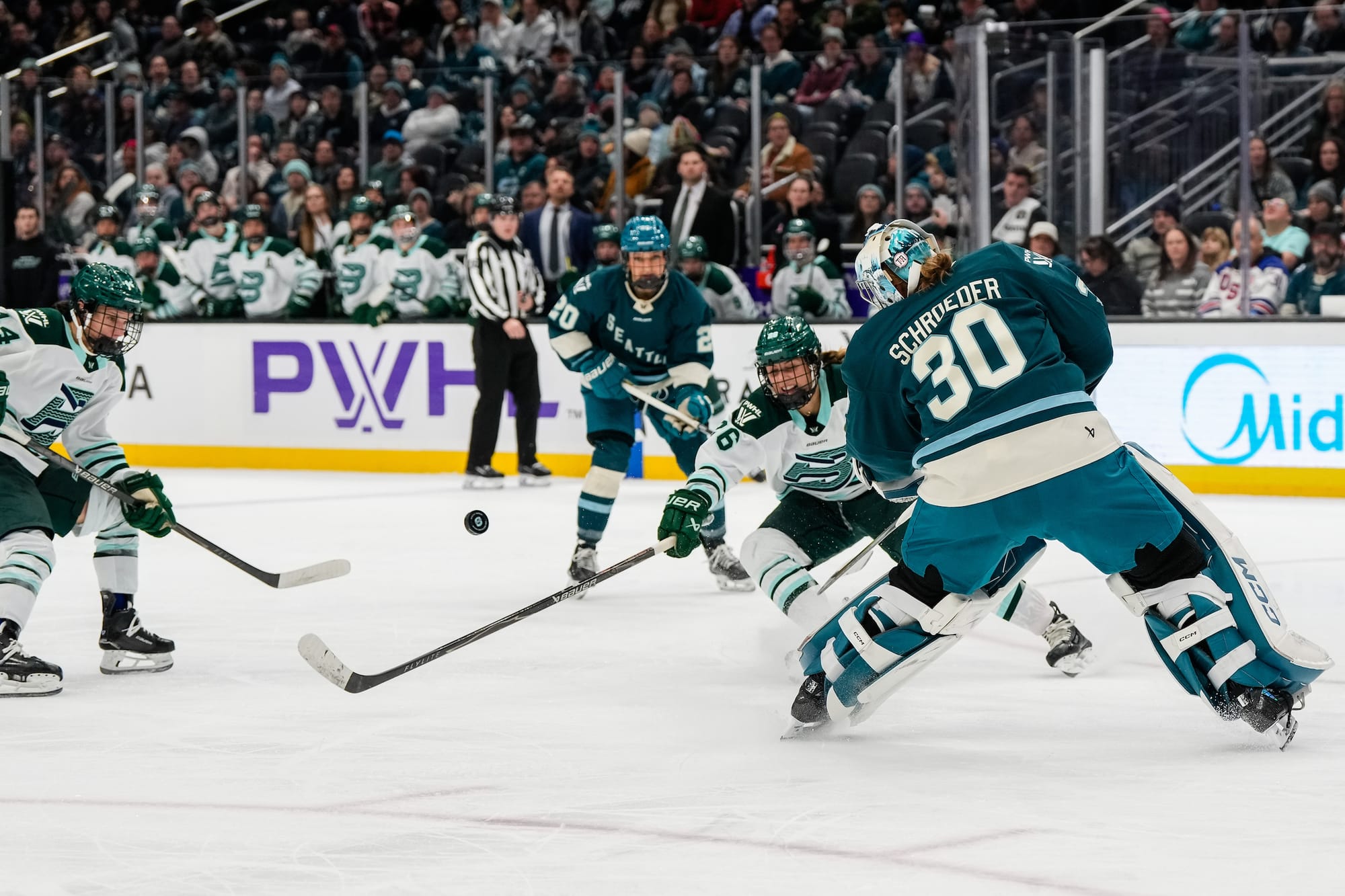 Schroeder has come up to the hash marks of the circles to send the puck back up ice and out of danger. There are Fleet players in the background trying to intercept it. Schroeder is wearing a teal home uniform, while the Fleet players are in white away ones. 