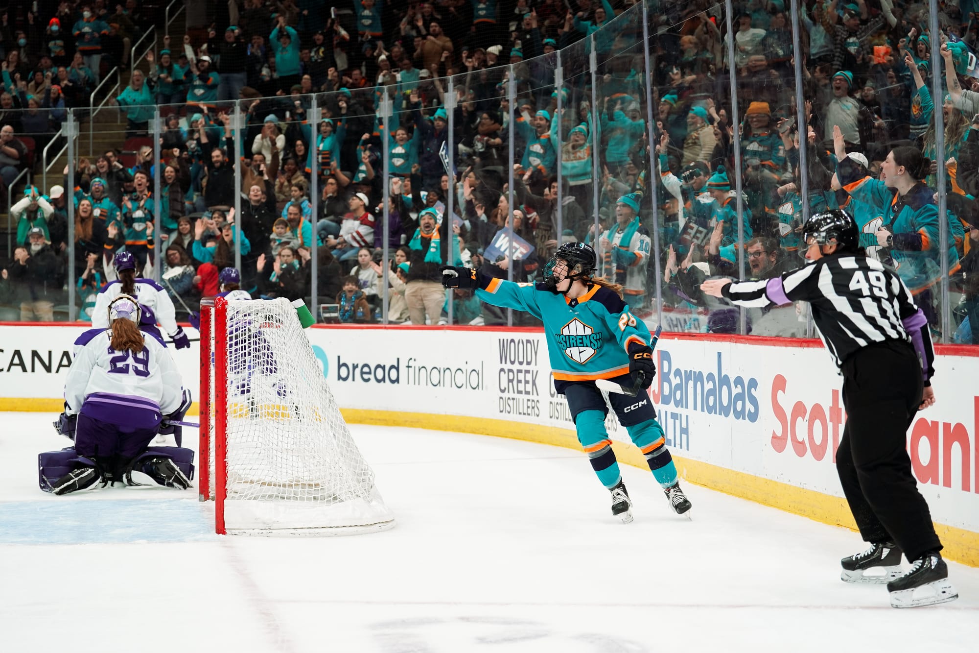 O'Brien smiles and points at the goal from behind the net in celebration, as the referee does the same and the crowd roars behind her. She is wearing a teal home uniform.