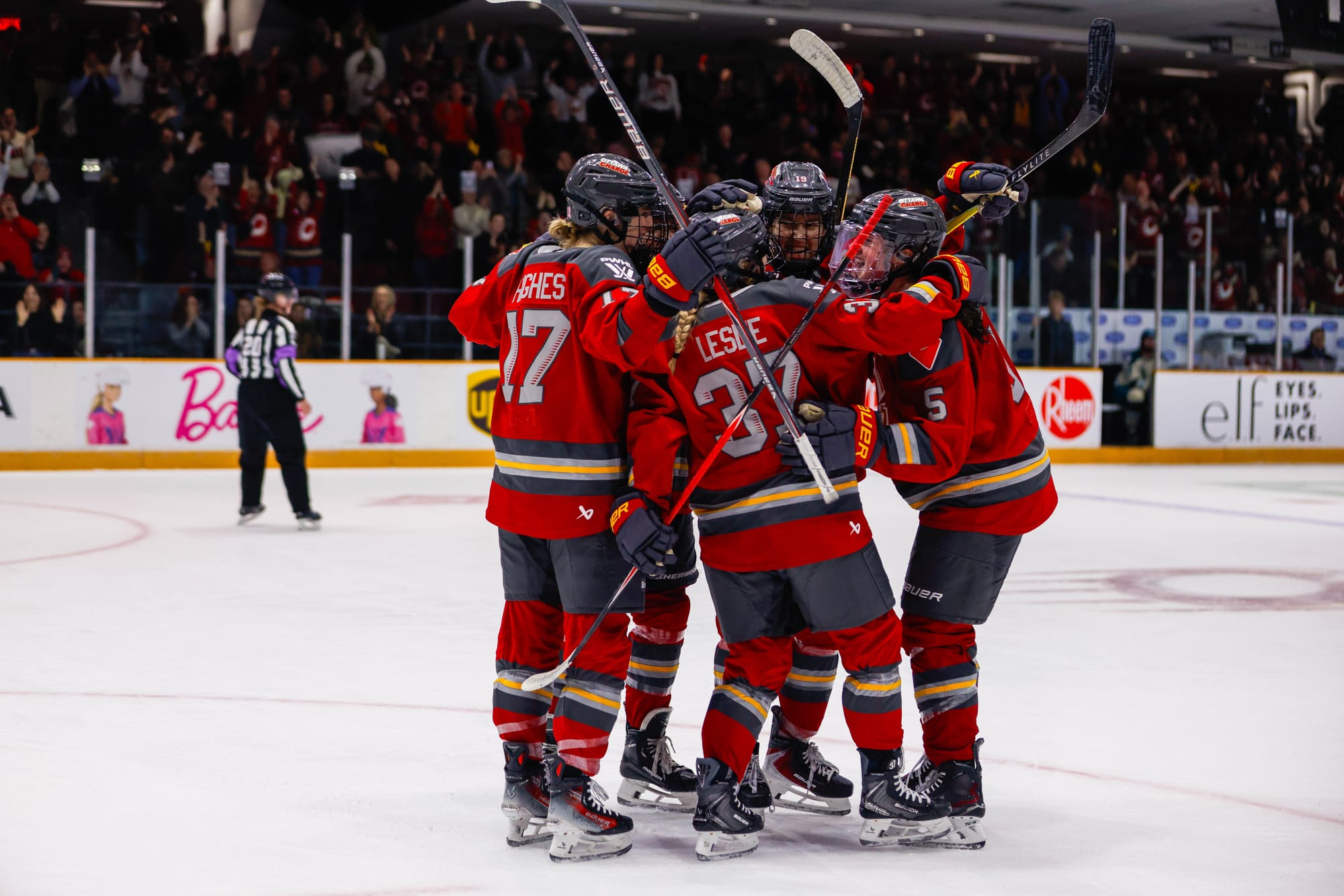 Charge players celebrate a goal with smiles and a tight group hug. They are all wearing red home uniforms.