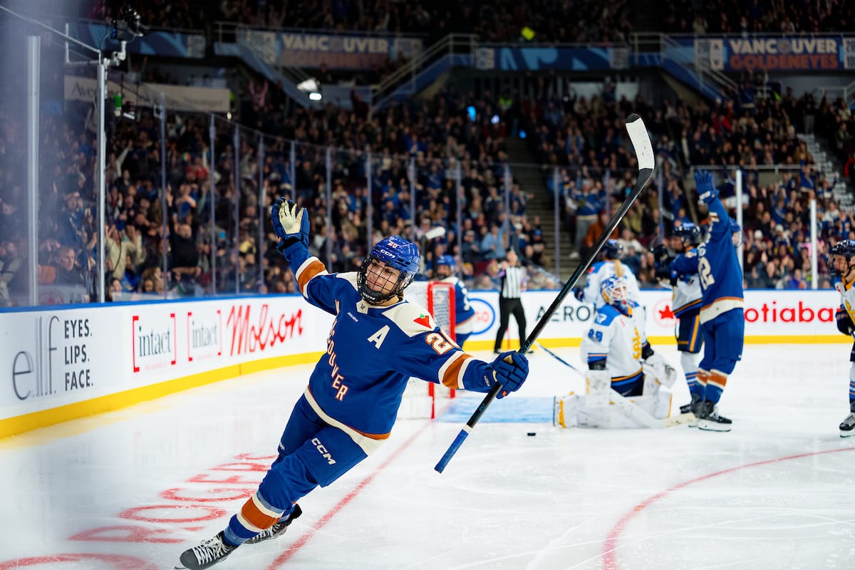 Nurse smirks and raises her arms as she skates away from the net, while the crowd roars in the background. She is wearing a blue home uniform.