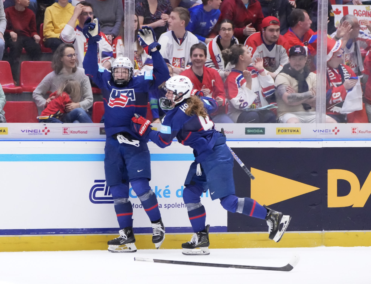 Janecke raises her arms after throwing her stick into the crowd and smiles as Harvey smiles and goes in for the hug. They are wearing blue home uniforms.