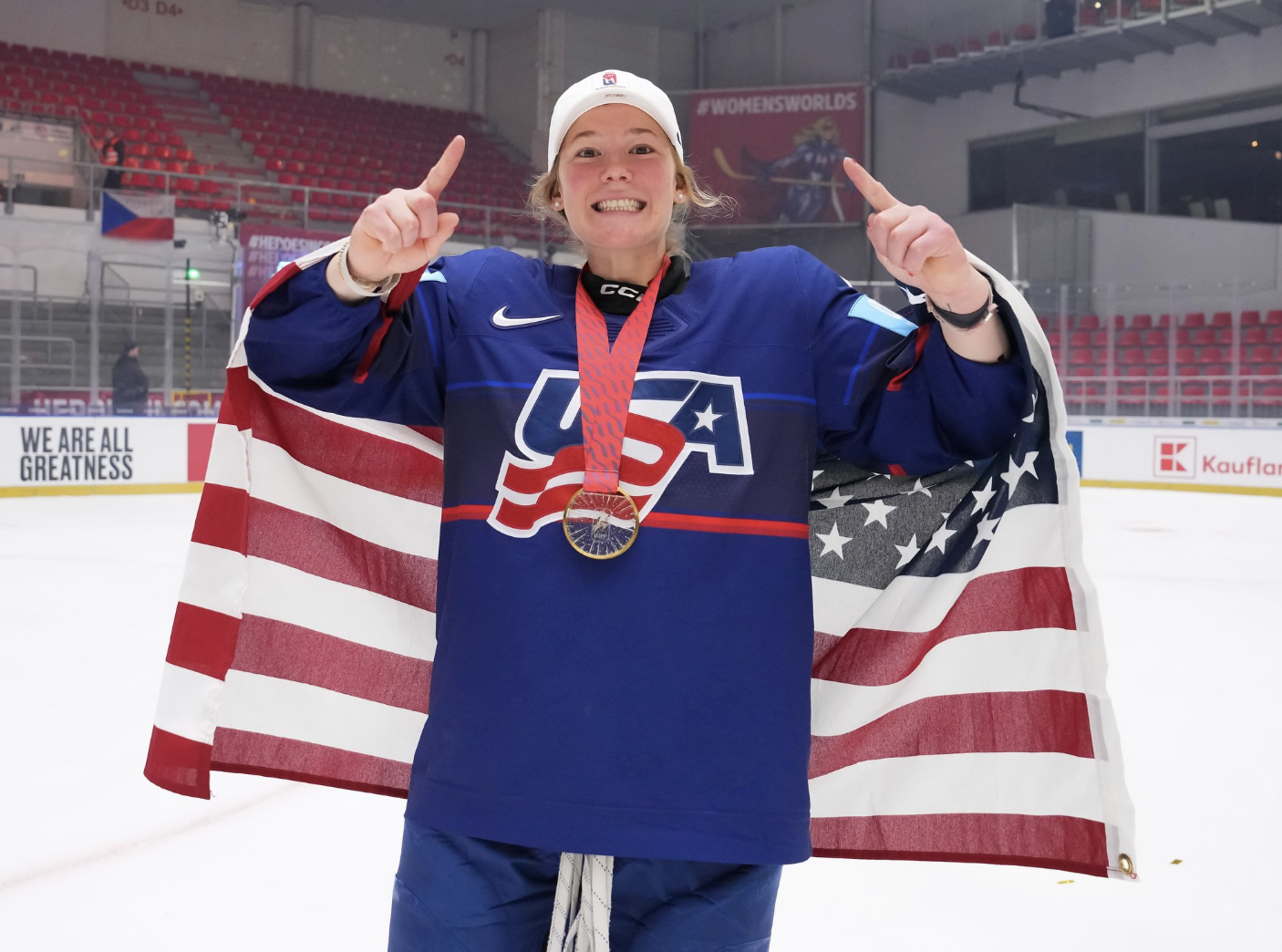 Winn smiles at the camera with a gold medal around her neck and an American flag wrapped around her back. She is holding up a number one with each hand, and wearing a blue home uniform.