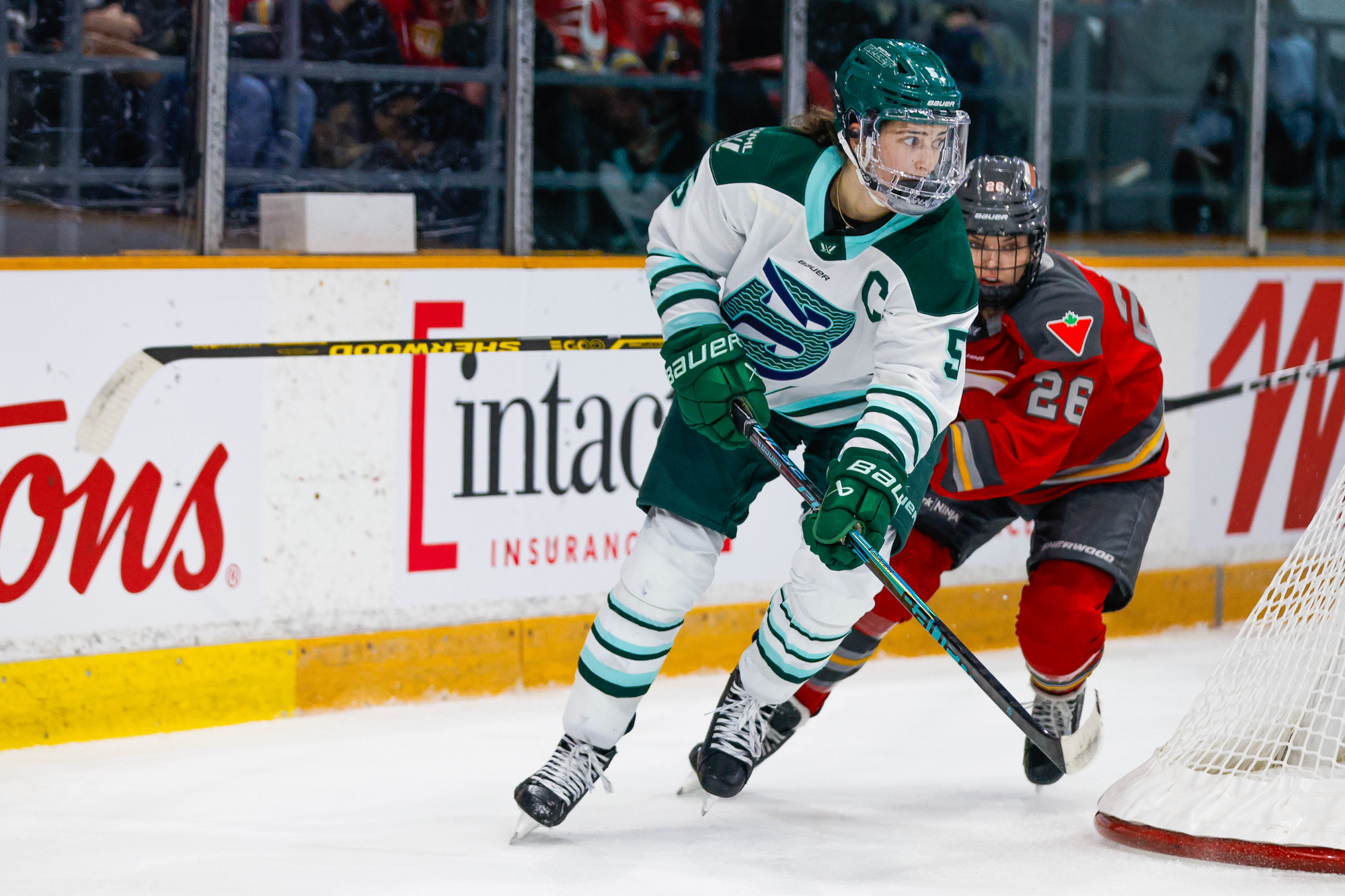 Keller skates around the back of the net, watching a play unfold out of frame. She is being chased by an Ottawa defender. Keller is wearing a white away uniform, while the Ottawa player is in a red home one.