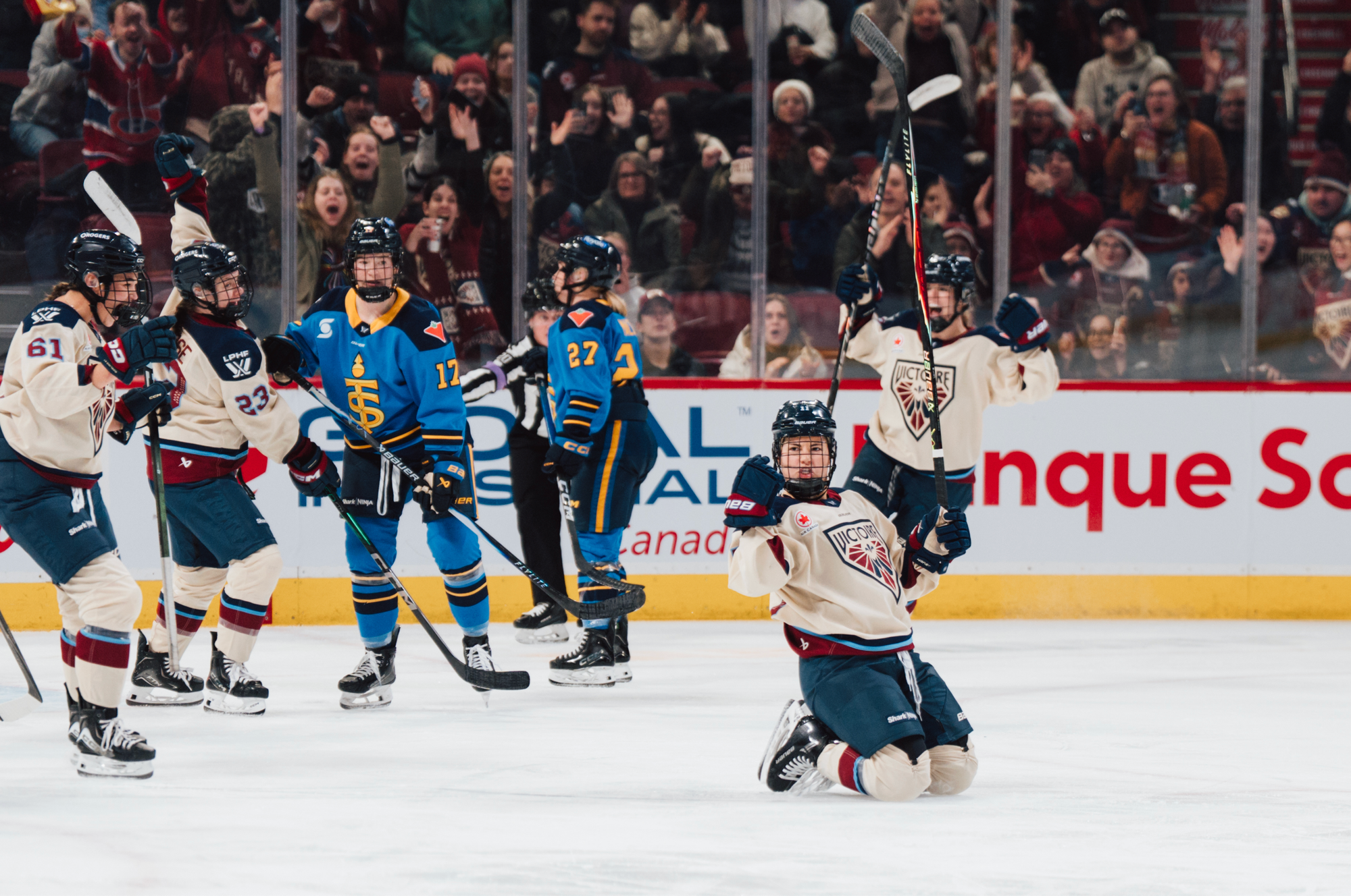 Roque yells and pumps her fists from her knees, while her teammates celebrate and skate towards her for a hug. They are all wearing cream away uniforms.