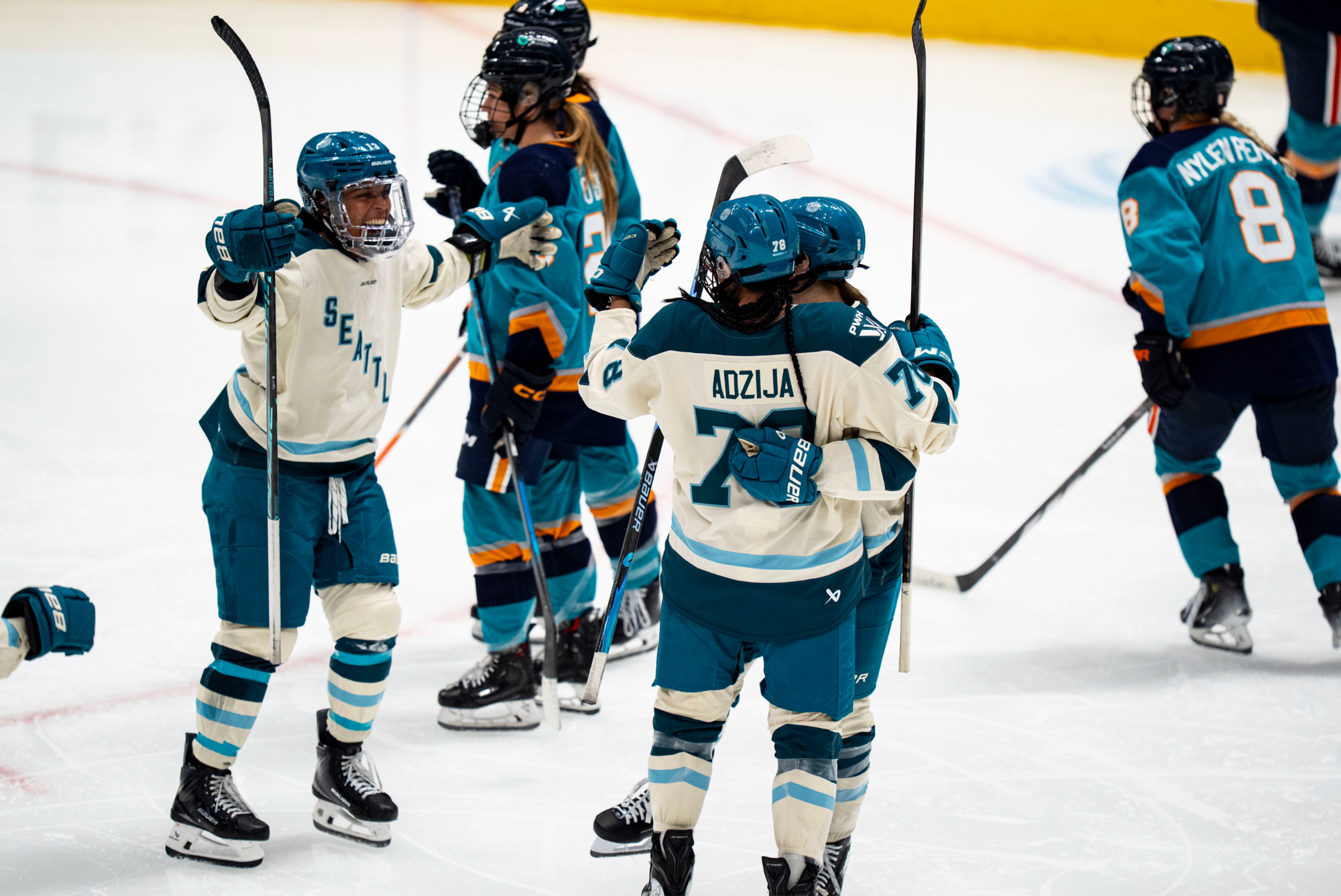 Grant-Mentis smiles and skates toward Adzija and another teammate, who are already hugging, with her arms open wide. They are all wearing cream away uniforms.