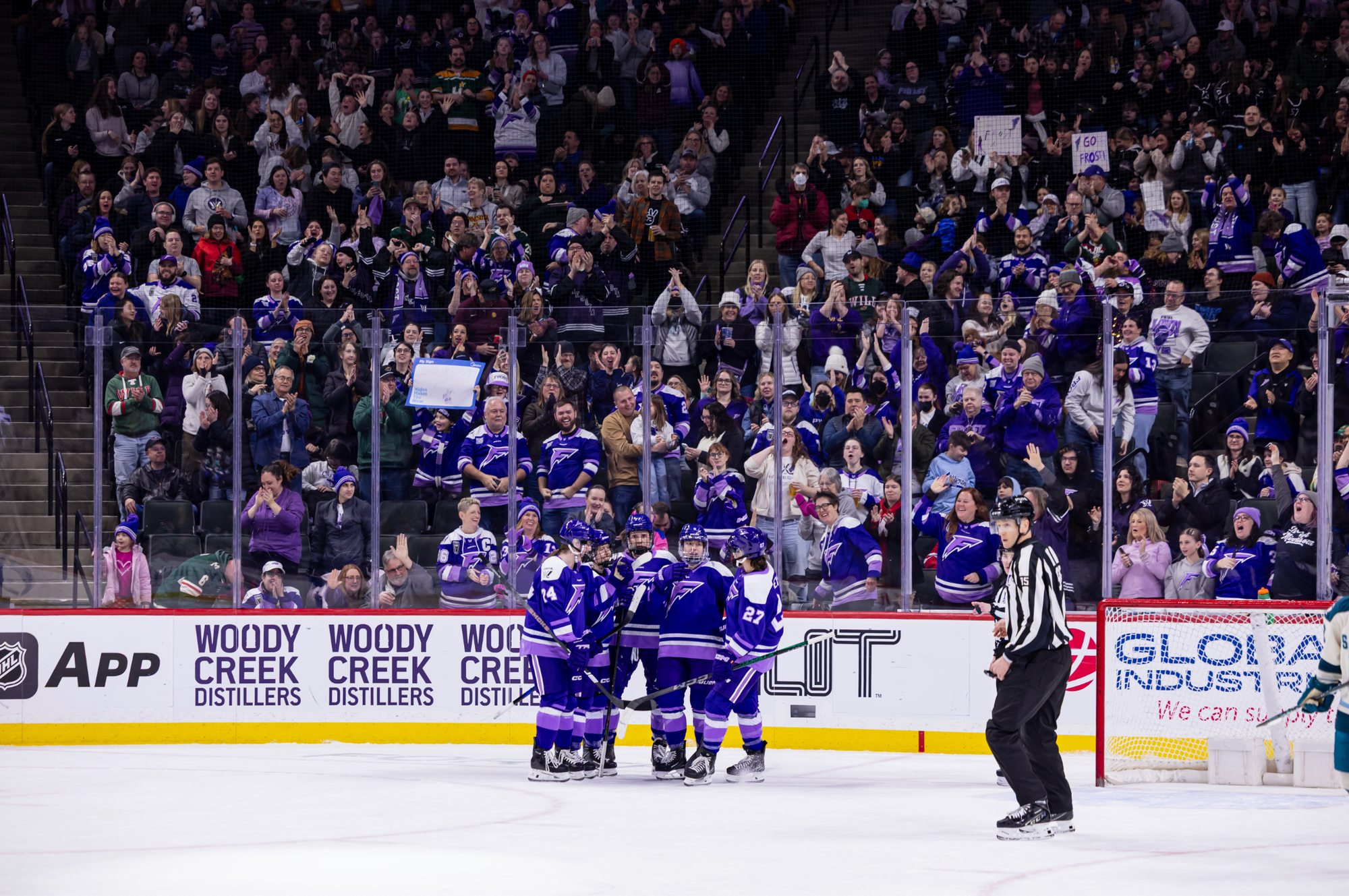 Minnesota players hug off to the side of the net following a goal. The crowd is on their feet, cheering them on in the background. The players are all wearing purple home uniforms.