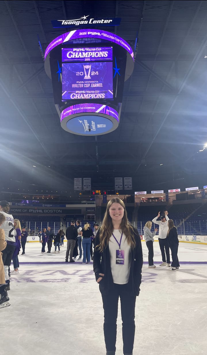 A photo of me on the ice postgame. I'm wearing a black sweater and pants with a white shirt, and my press pass.