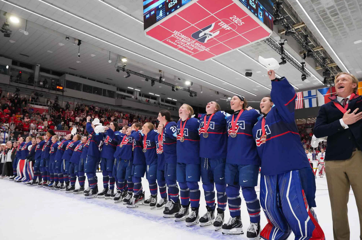 Team USA stands arm in arm on the blueline, belting along with their anthem as it plays on the speakers. They are wearing blue uniforms.