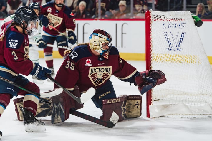 Desbiens looks behind the net as she prepares to clamp down on the puck when it pops back out. She is wearing a maroon home uniform.