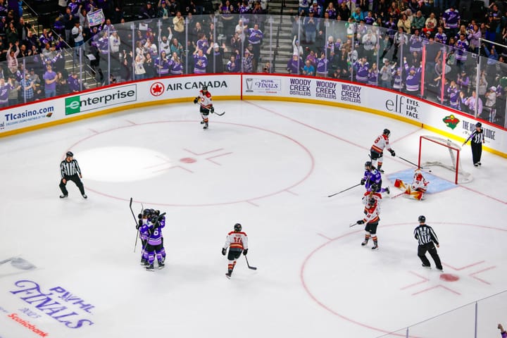 An above-ice shot of Frost players celebrating with a tight group hug while Ottawa players look disappointed. The Frost are in purple, while the Charge are in white.