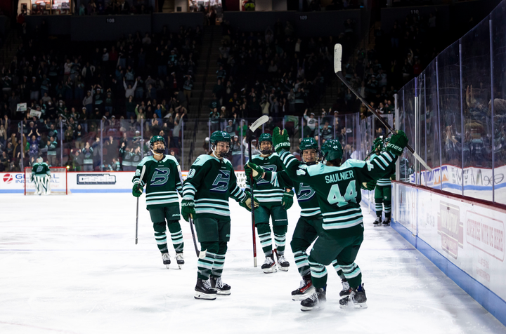 Saulnier raises her arms in celebration as she skates toward her teammates in celebration. They are all wearing green home uniforms.