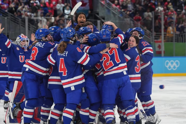 Team USA celebrates their gold medal win with smiles and a big, tight group hug at center ice. They are all wearing blue home uniforms.